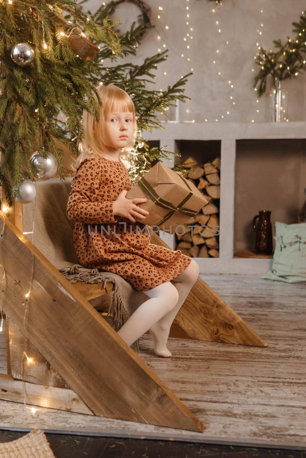 A little blonde girl is sitting on a wooden staircase in a Scandinavian interior decorated in a New Year's style. A child holds a Christmas gift in a craft package.
