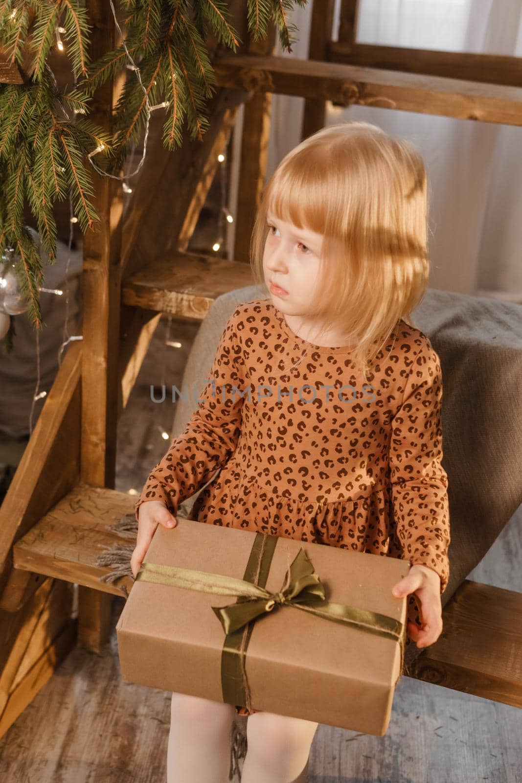 A little blonde girl is sitting on a wooden staircase in a Scandinavian interior decorated in a New Year's style. A child holds a Christmas gift in a craft package.