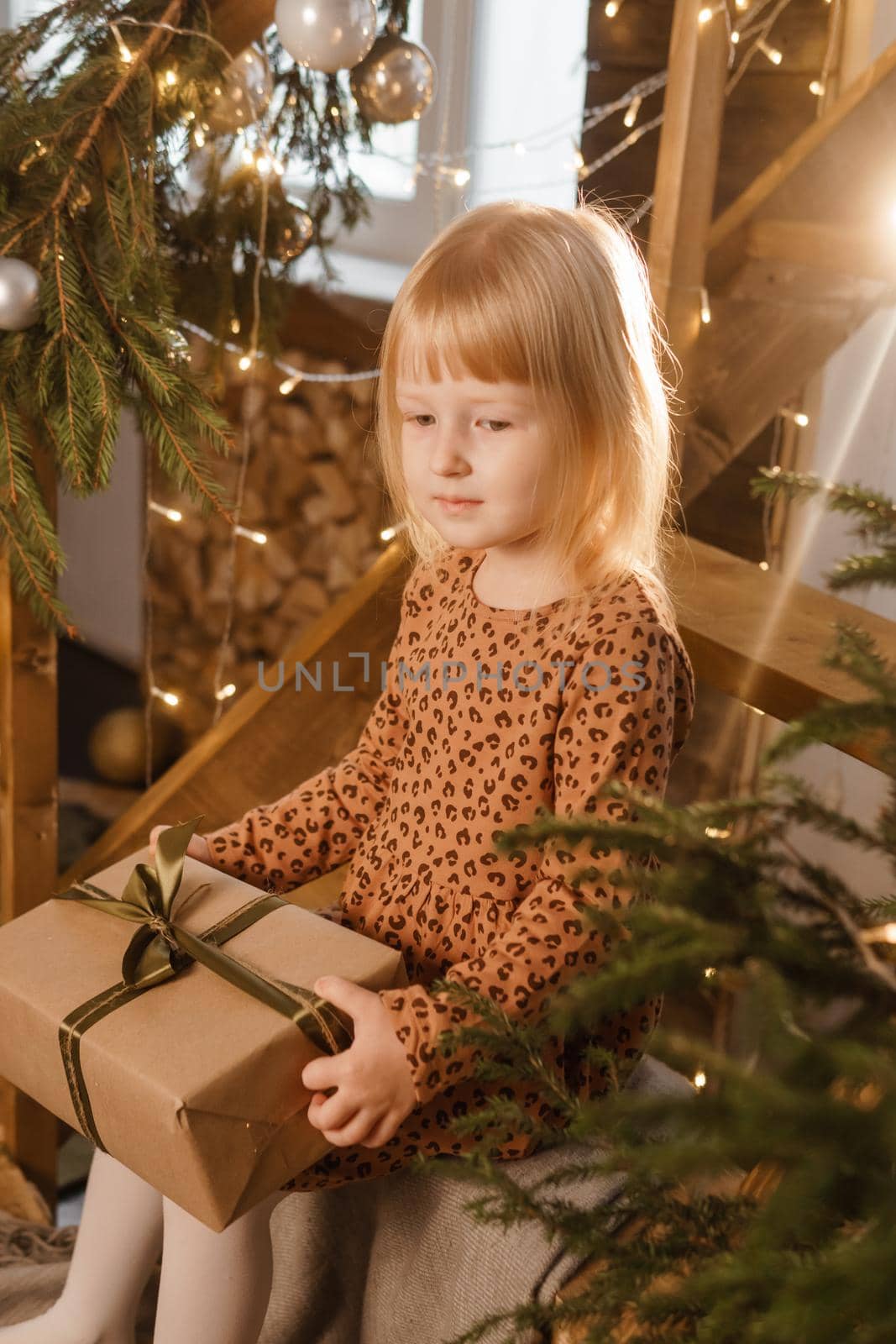 A little blonde girl is sitting on a wooden staircase in a Scandinavian interior decorated in a New Year's style. A child holds a Christmas gift in a craft package.