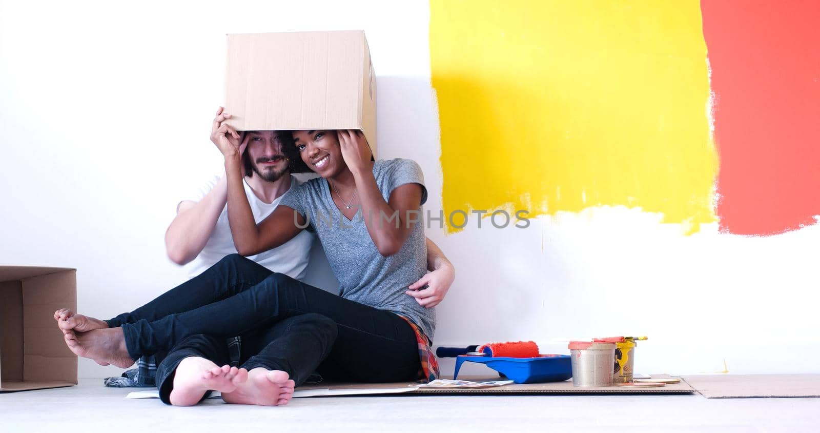 Happy young multiethnic couple relaxing and playing with cardboard boxes after painting a room in their new house on the floor