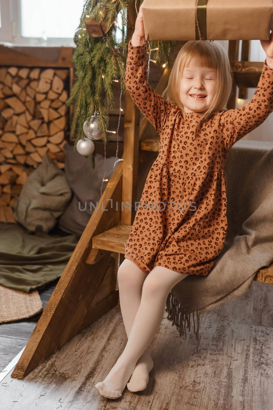 A little blonde girl is sitting on a wooden staircase in a Scandinavian interior decorated in a New Year's style. A child holds a Christmas gift in a craft package.