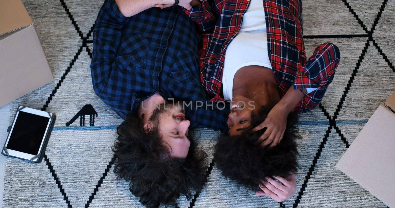 Top view of attractive young multiethnic couple moving in, holding hands, looking at camera and smiling while lying among cardboard boxes