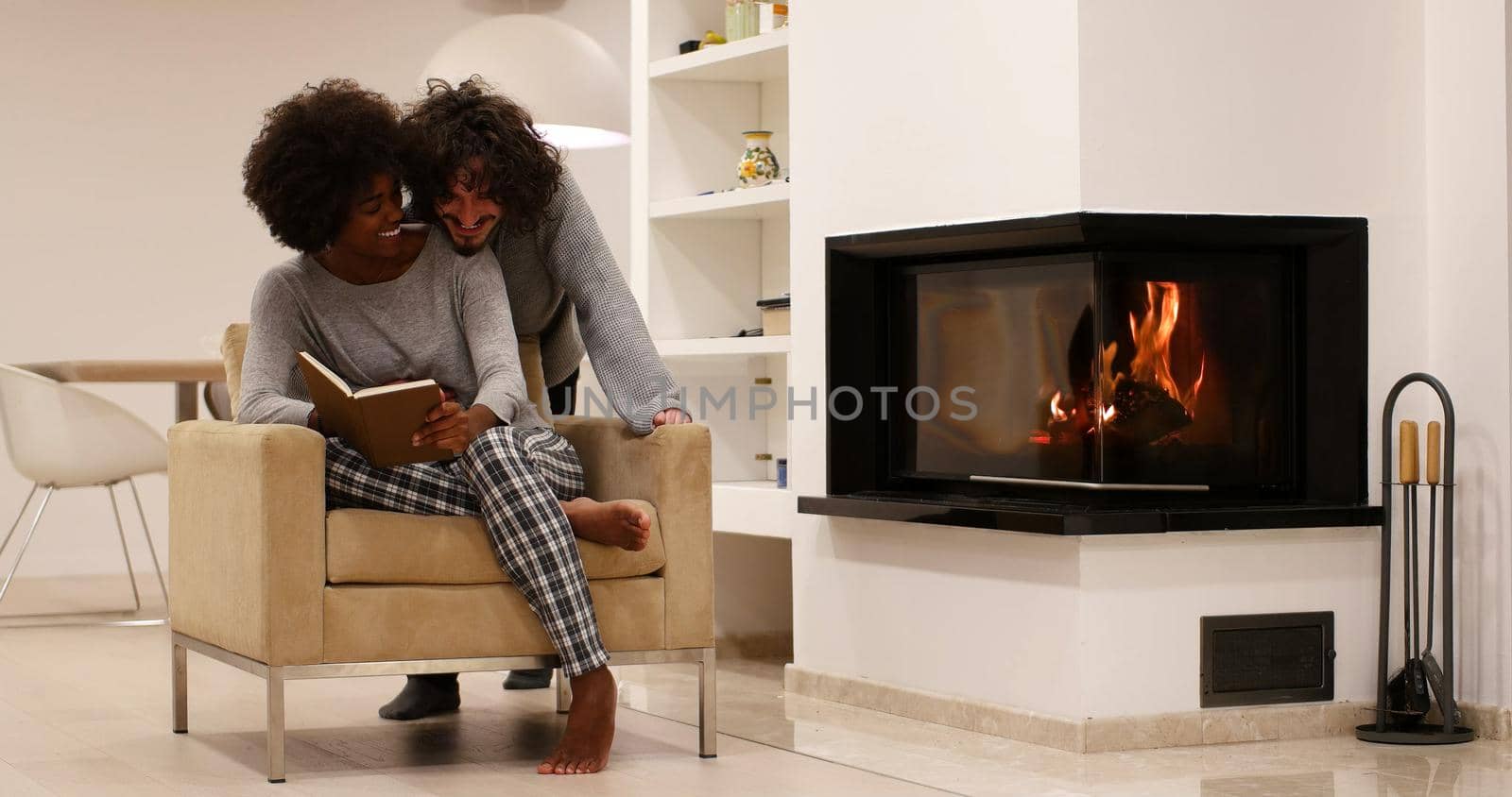 Young beautiful multiethnic couple hugging in front of fireplace at home when reading a book at autumn day