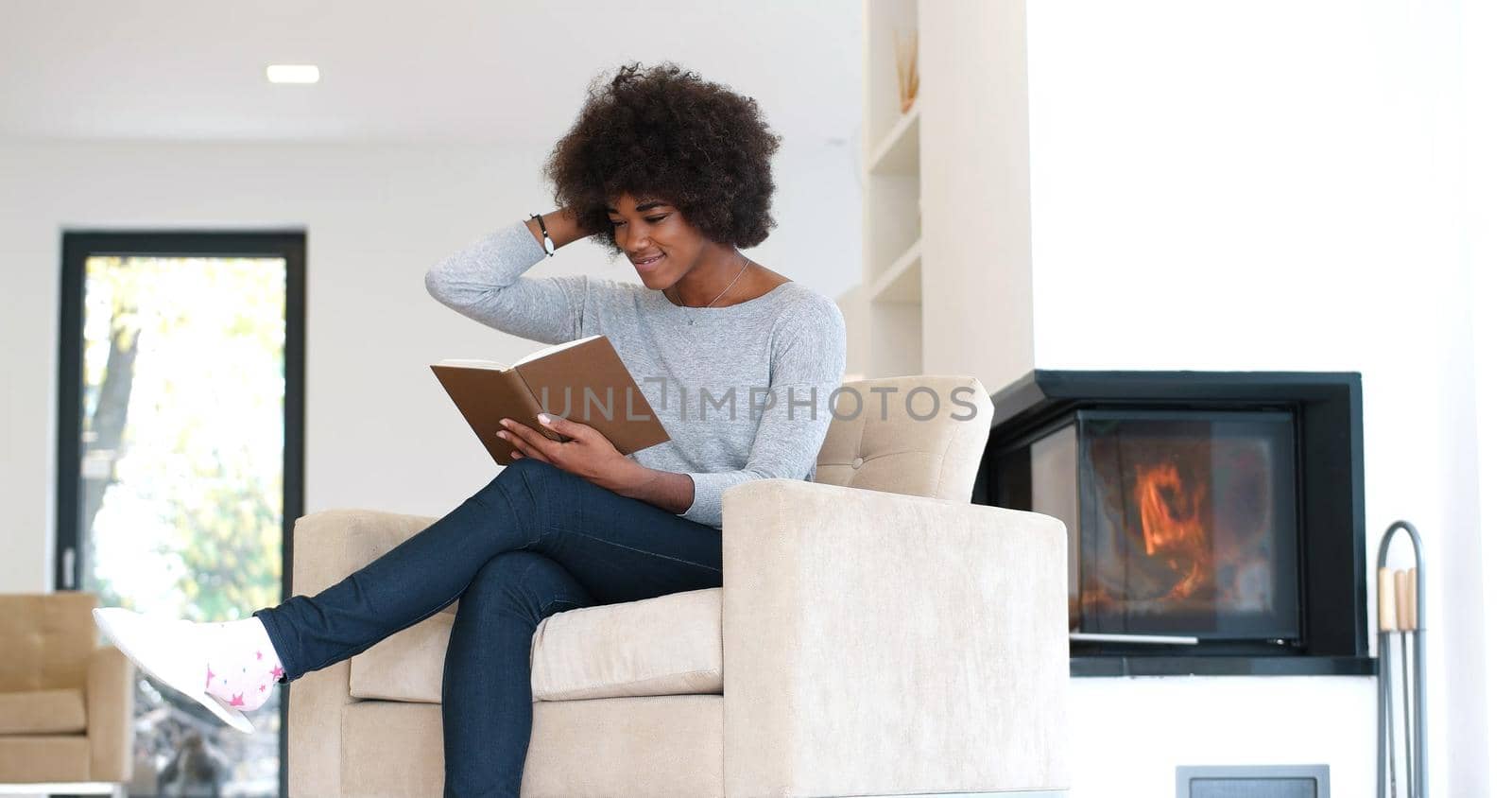 Young beautiful african american woman sitting in front of fireplace at home on a cold autumn day and reading book