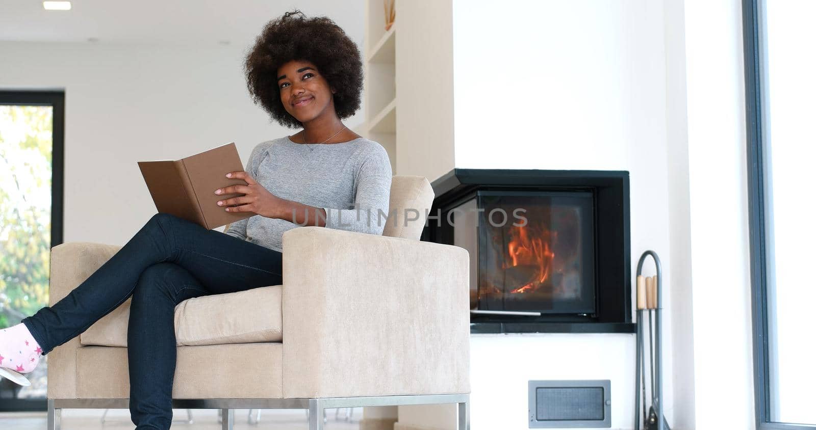 Young beautiful african american woman sitting in front of fireplace at home on a cold autumn day and reading book