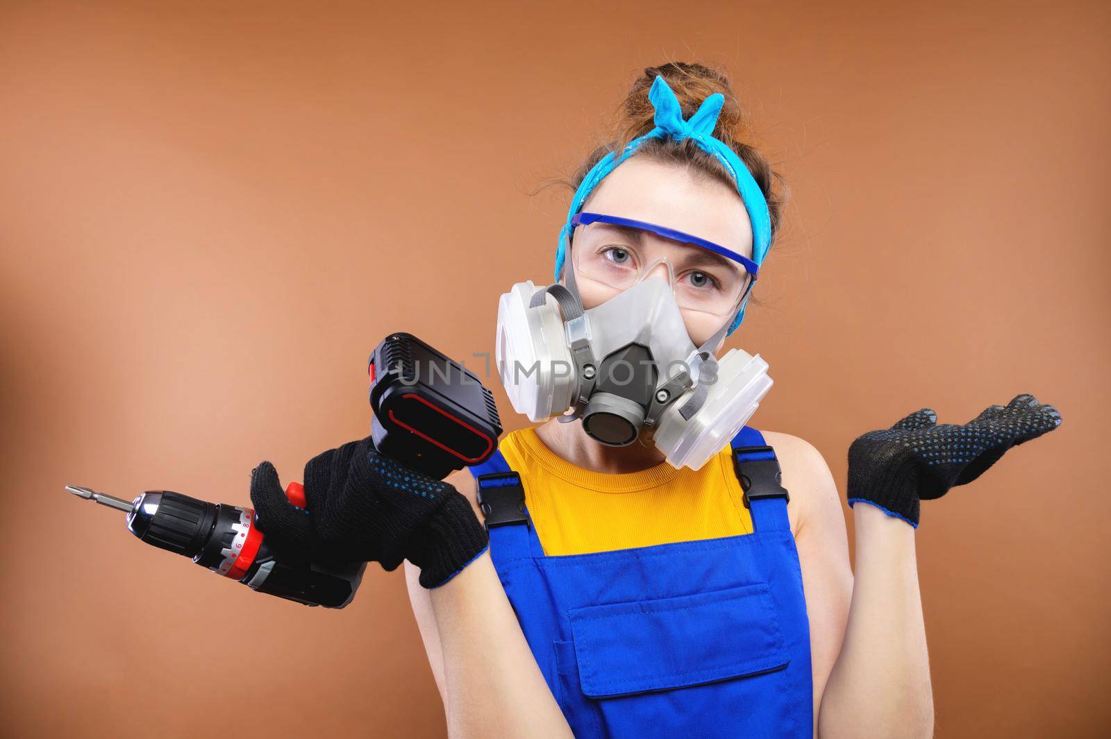 Portrait of a caucasian young woman in the uniform of a superintendent of streetwear in overalls, respirator and goggles with a screwdriver in hand. Looks into the camera. In annoyance, he throws up his hands after a mistake or misunderstanding. Builder bug concept