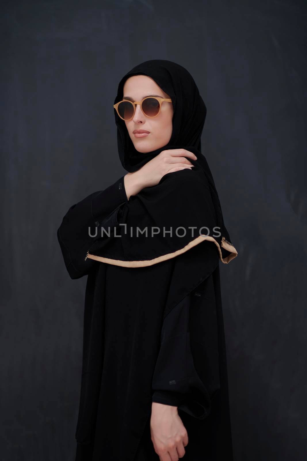 Young muslim in traditional clothes or abaya and sunglasses posing in front of black chalkboard. Arab woman representing modern arabic lifestyle, islamic  fashion and Ramadan kareem concept