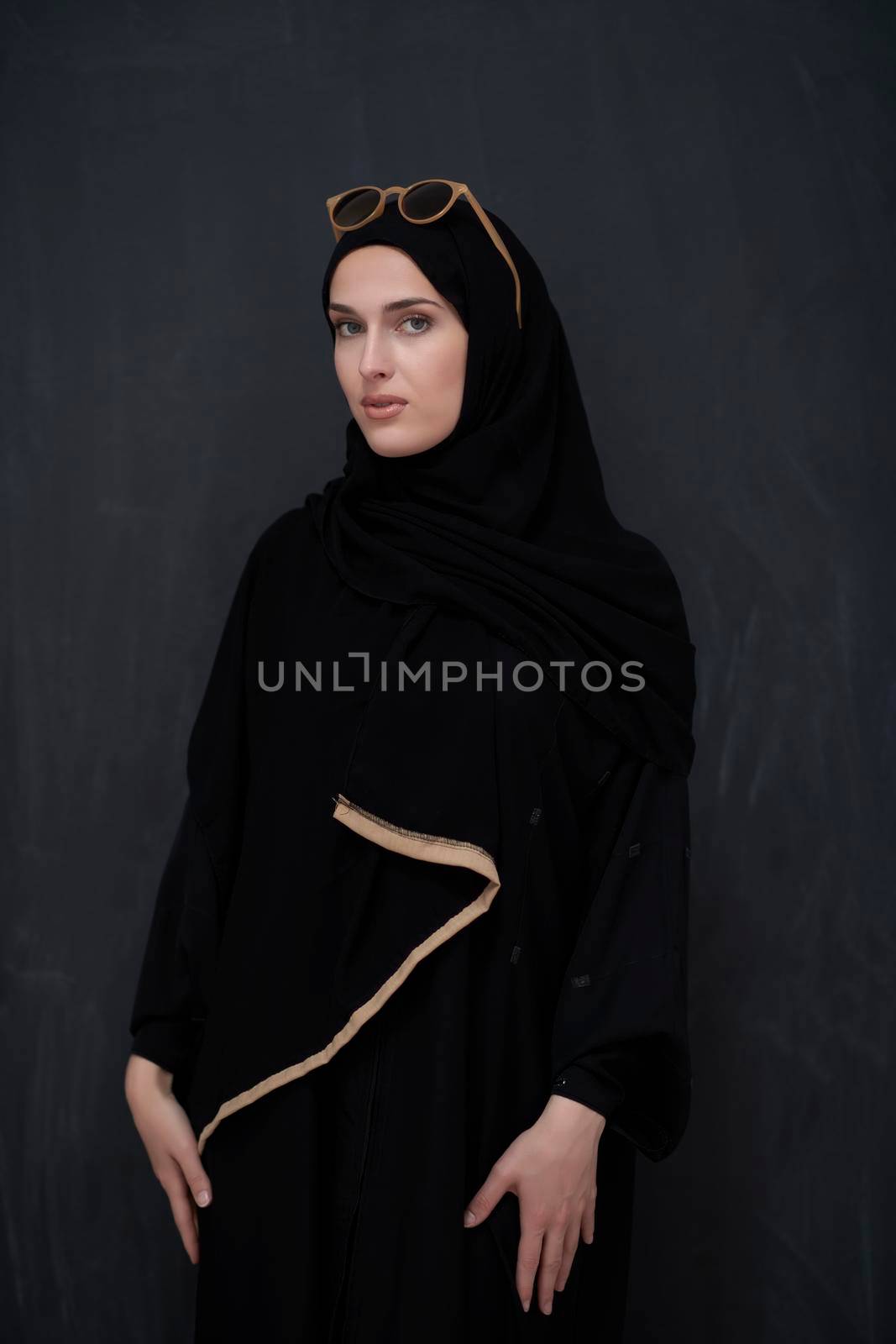 Young muslim in traditional clothes or abaya and sunglasses posing in front of black chalkboard by dotshock