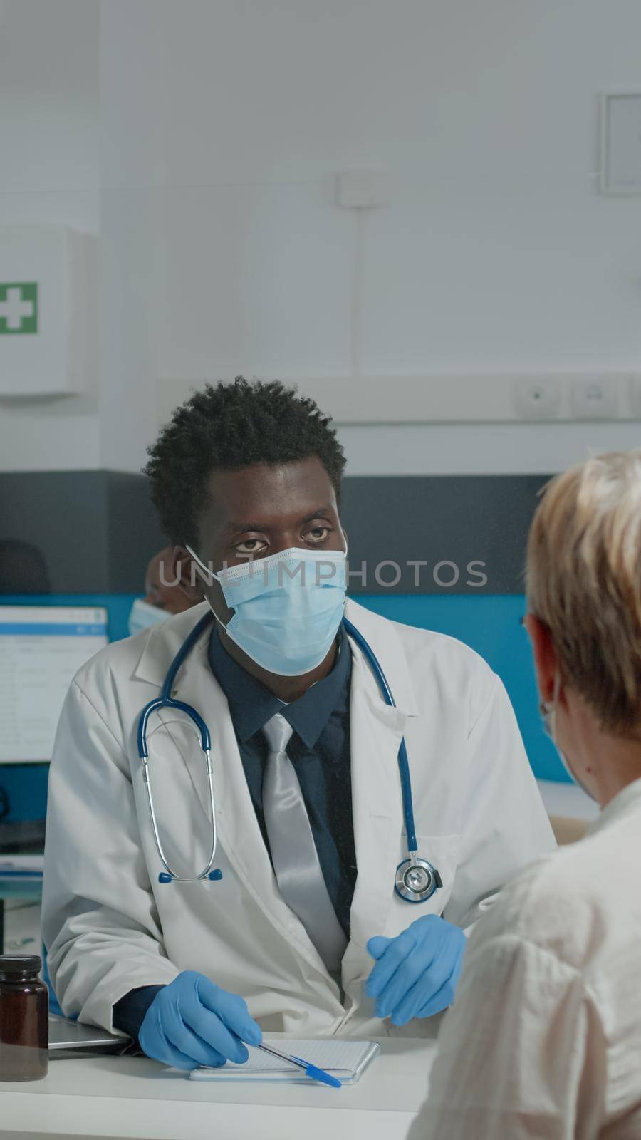 Medic of african american ethnicity with face mask consulting elder woman behind plexiglass wall. Black doctor doing annual healthcare checkup with old patient during pandemic in cabinet