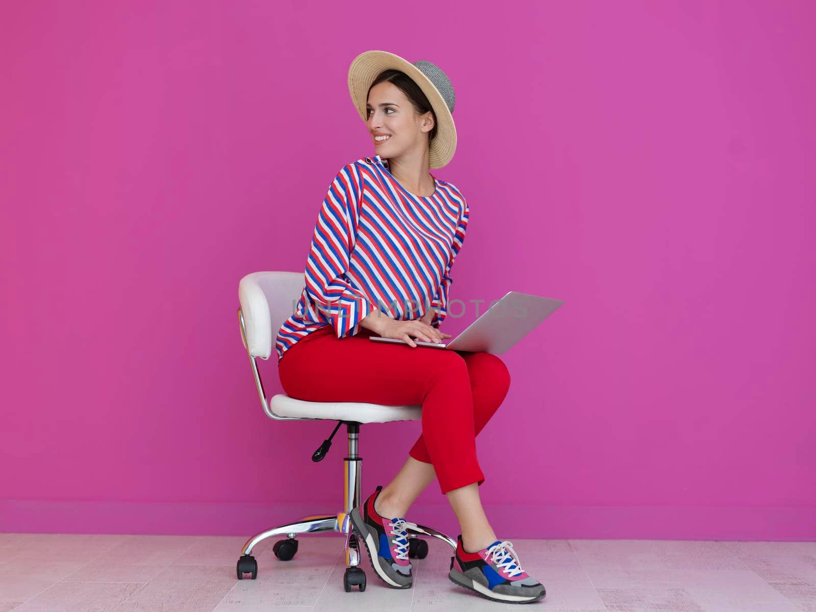 Portrait of young woman sitting on the chair and holding laptop on the lap isolated on pink background. Female model presenting fashion and technologz concept