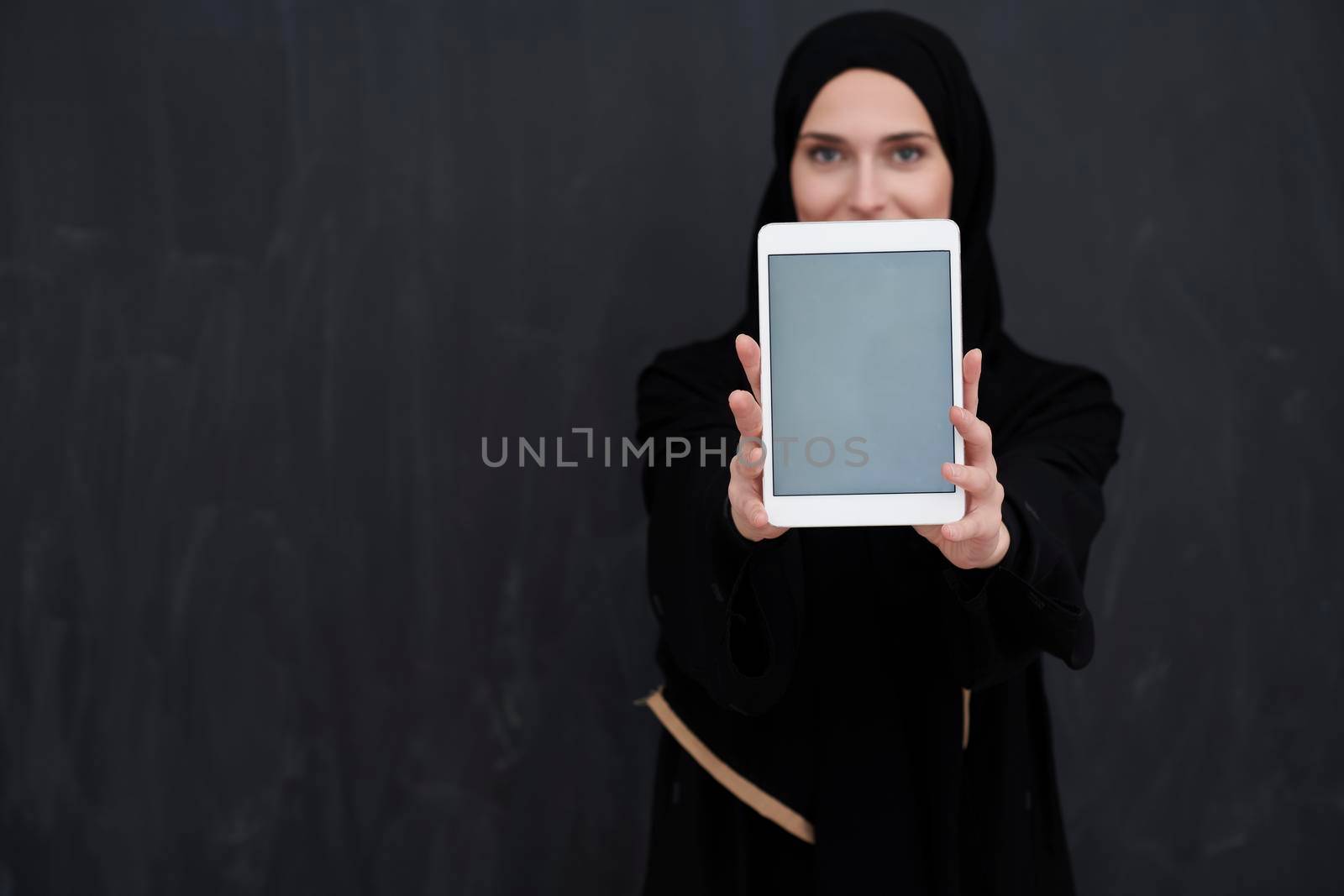 Young Arab businesswoman in traditional clothes or abaya and glasses holding tablet computer in front of black chalkboard representing modern islam fashion and technology