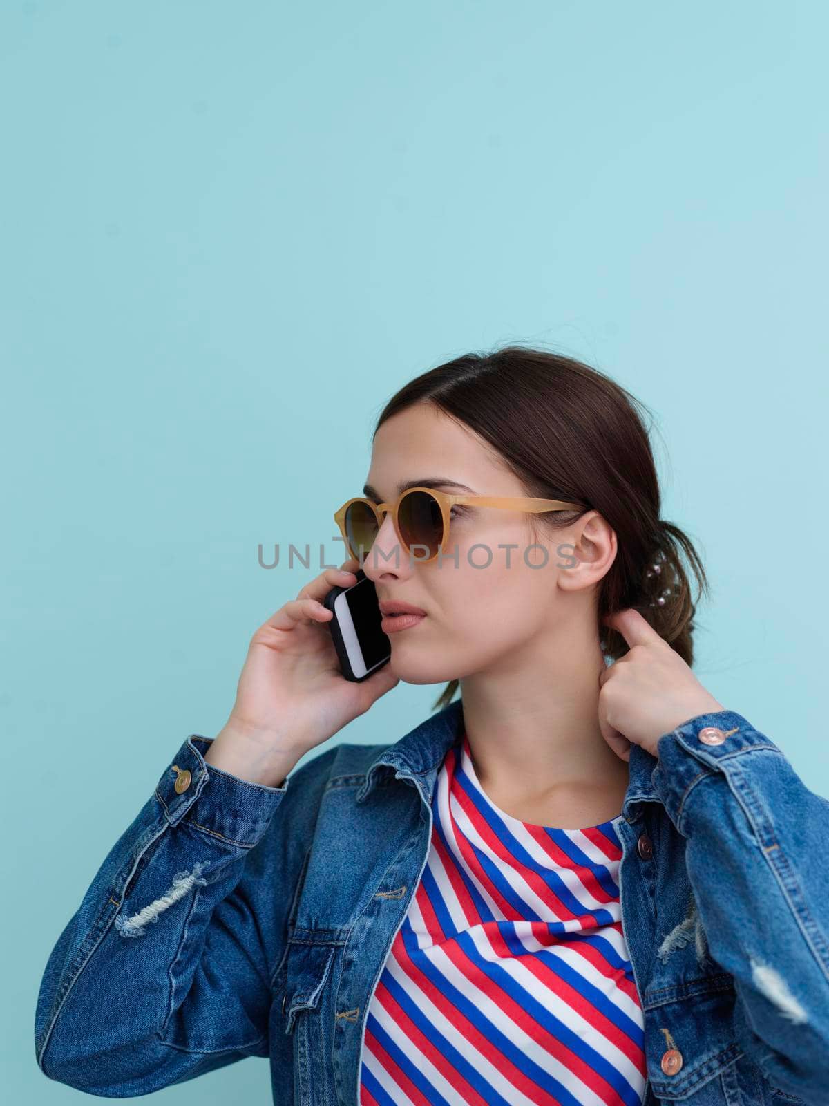 Portrait of young girl talking on the phone while standing in front of blue background. Female model wearing sunglasses representing modern fashion and technology concept