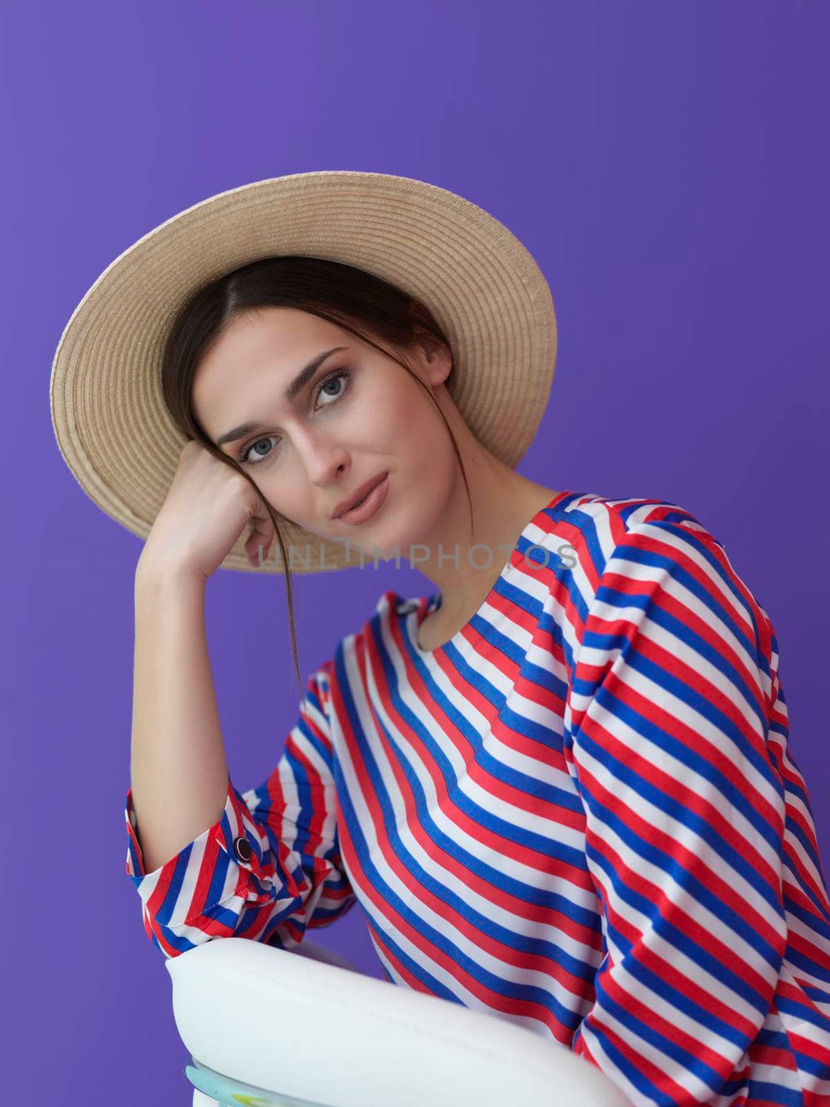 Portrait of young woman sitting on the chair isolated on purple background. Female model in casual and modern clothes posing in the studio