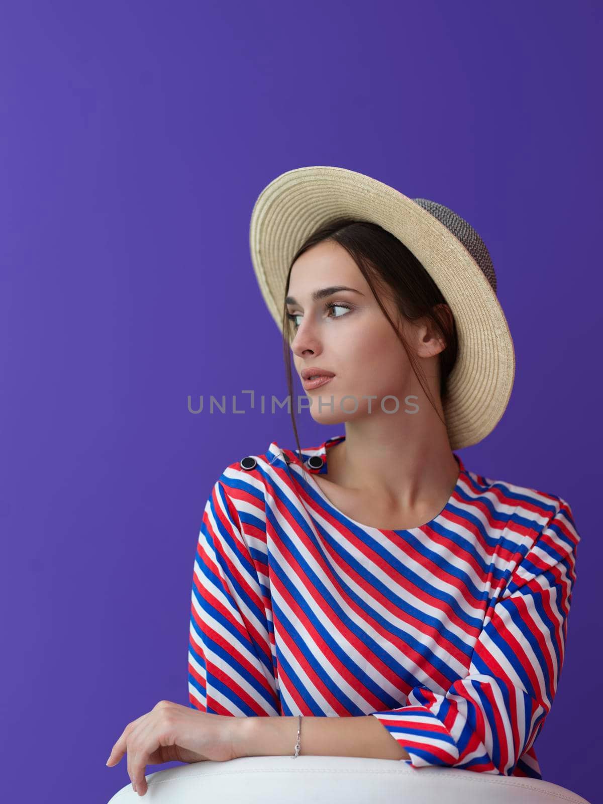 Portrait of young woman sitting on the chair isolated on purple background. Female model in casual and modern clothes posing in the studio