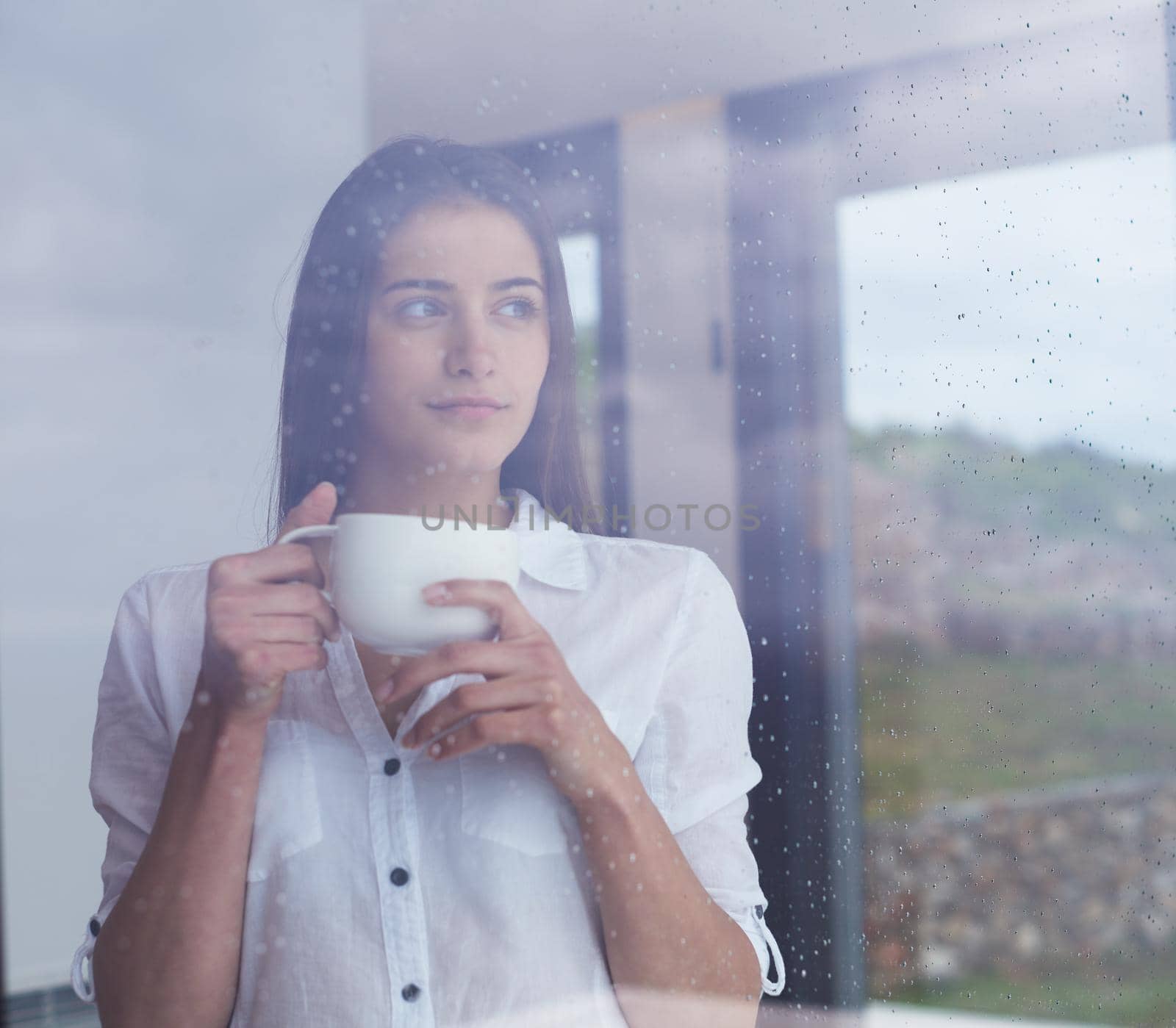 beautiful young woman drink first morning coffee at modern home interior with rain drops on big window door glass