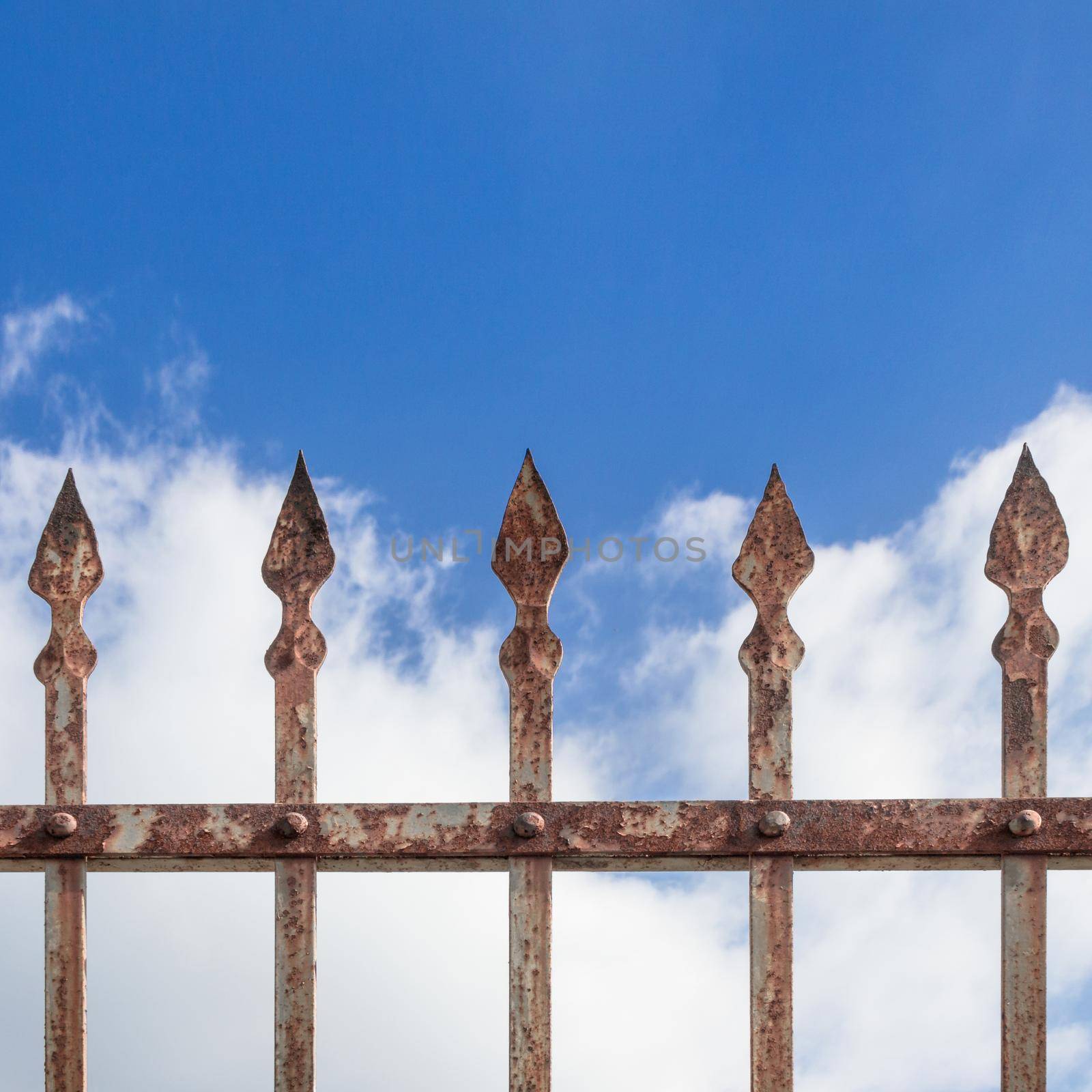 Details of a rusty fence decorated with spearheads and sky and clouds in the background.