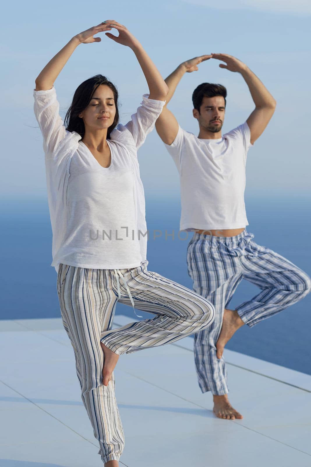 young couple practicing yoga at sunset in modern home terace with ocean and sunset in background