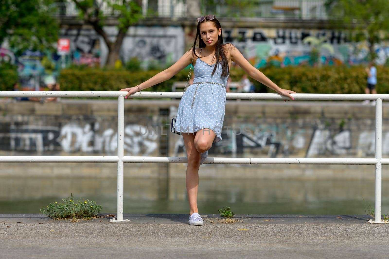 A portrait of a pretty young woman, taken in the summer in a city, leaning against a railing on a nearby river.