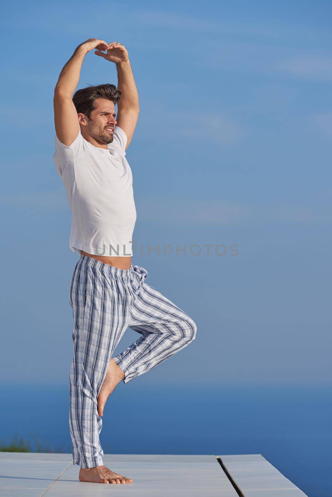 handsome young man practicing yoga on in modern home terace with ocean and sunset in background