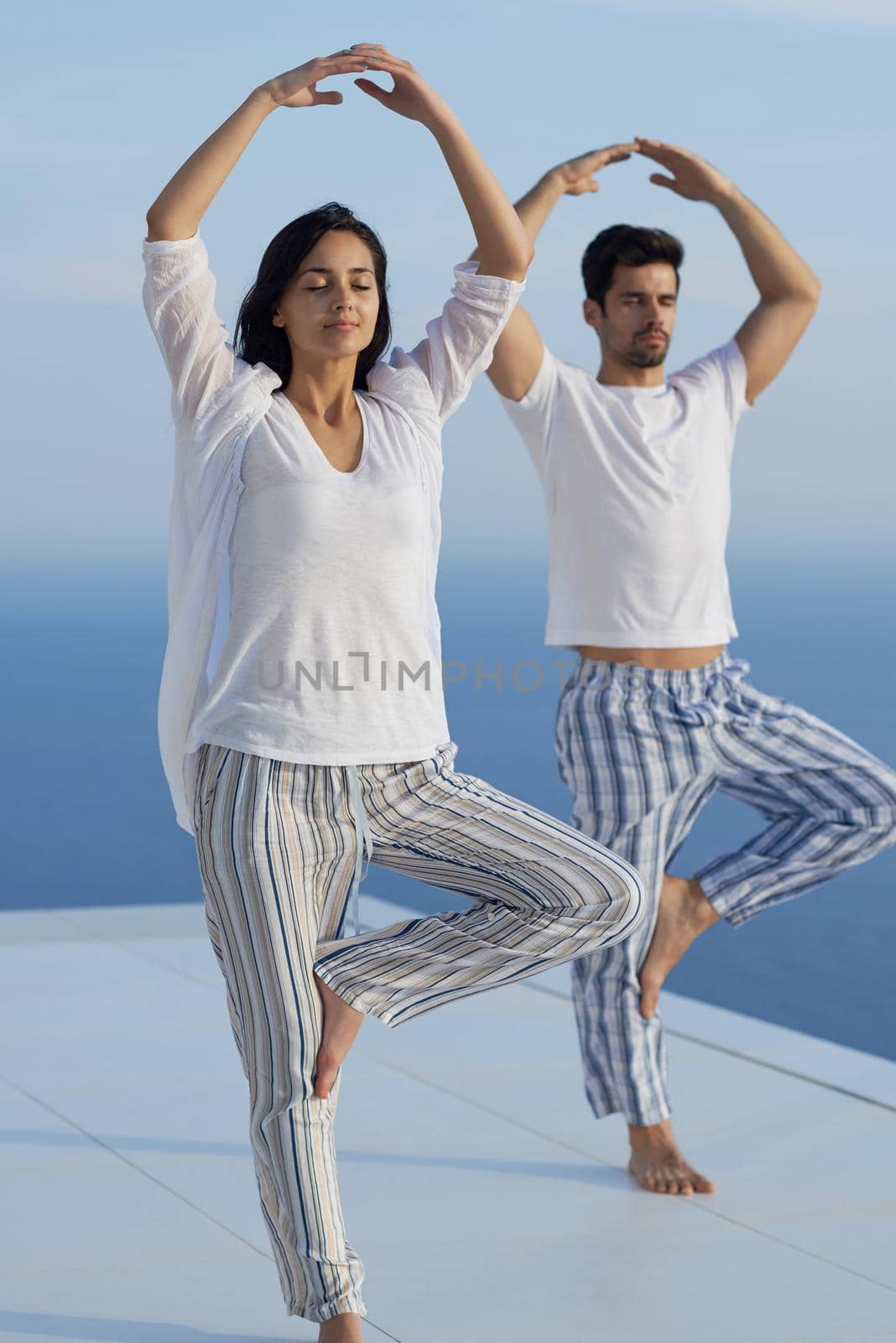 young couple practicing yoga at sunset in modern home terace with ocean and sunset in background