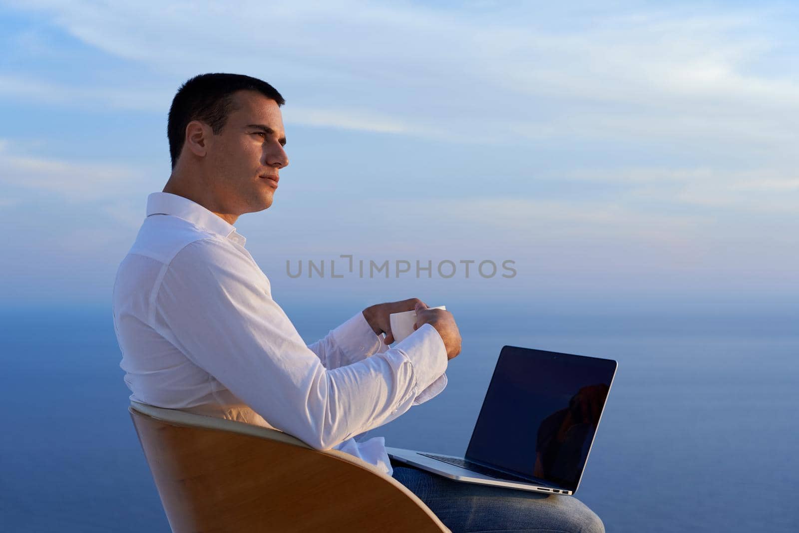 handsome young man relaxing and working on laptop computer at home balcony while looking sunset