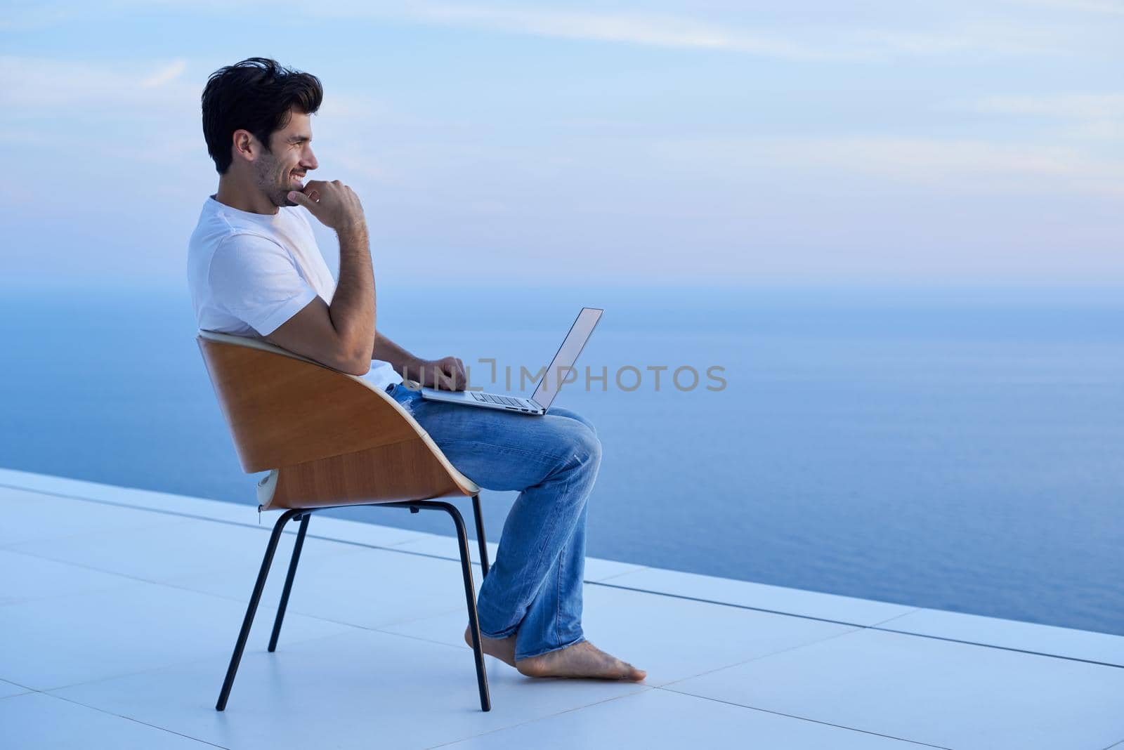 handsome young man relaxing and working on laptop computer at home balcony while looking sunset