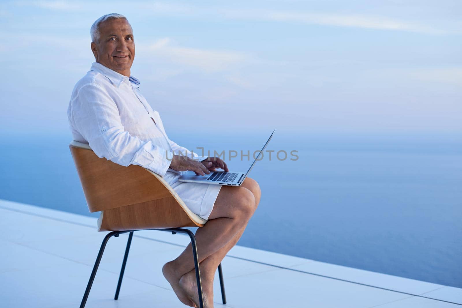 senior man working on laptop computer and looking ocean view sunset on balcony of modern home