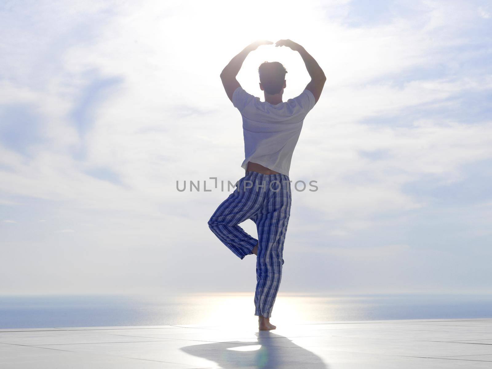 handsome young man practicing yoga on in modern home terace with ocean and sunset in background