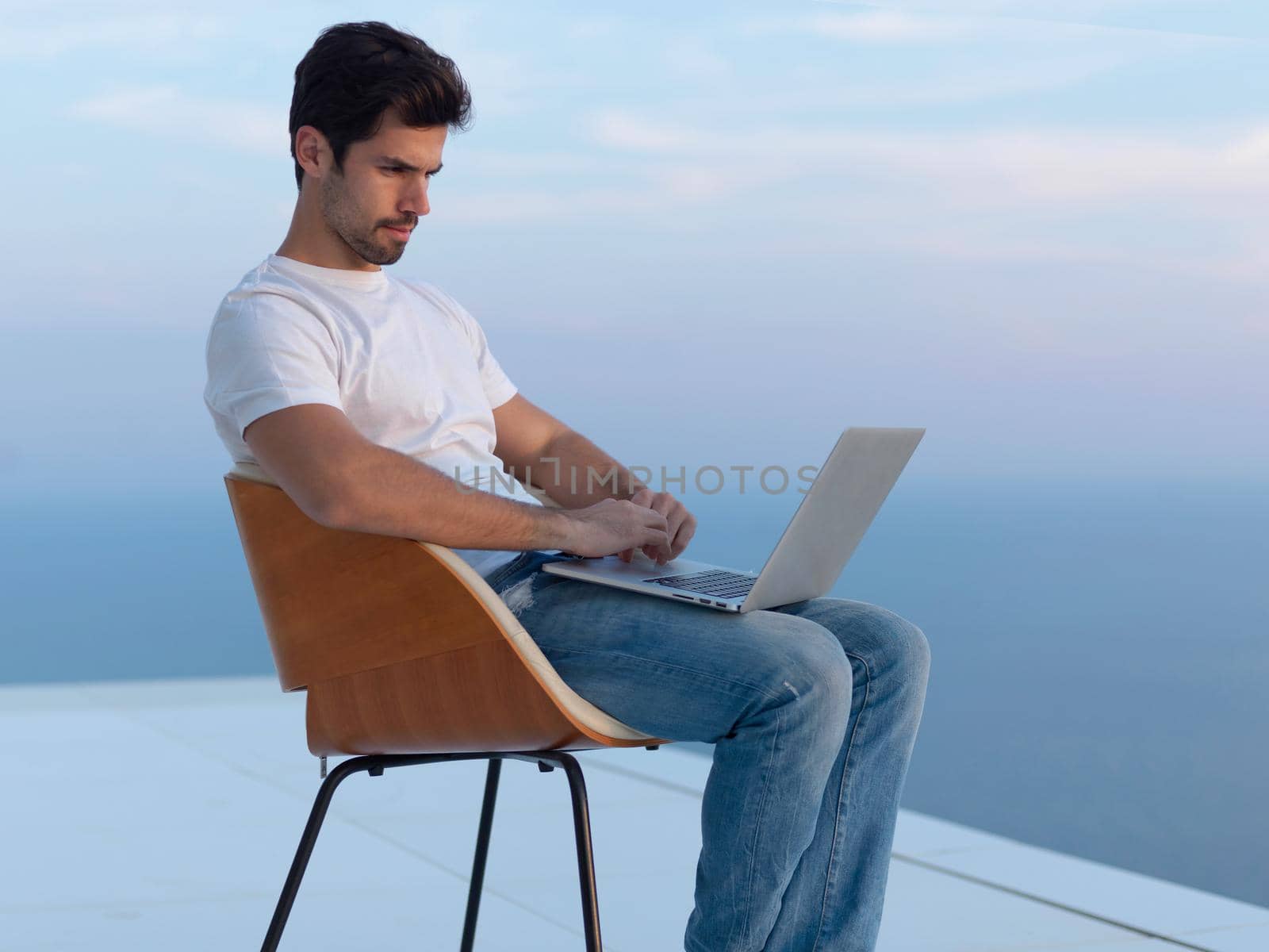 handsome young man relaxing and working on laptop computer at home balcony while looking sunset