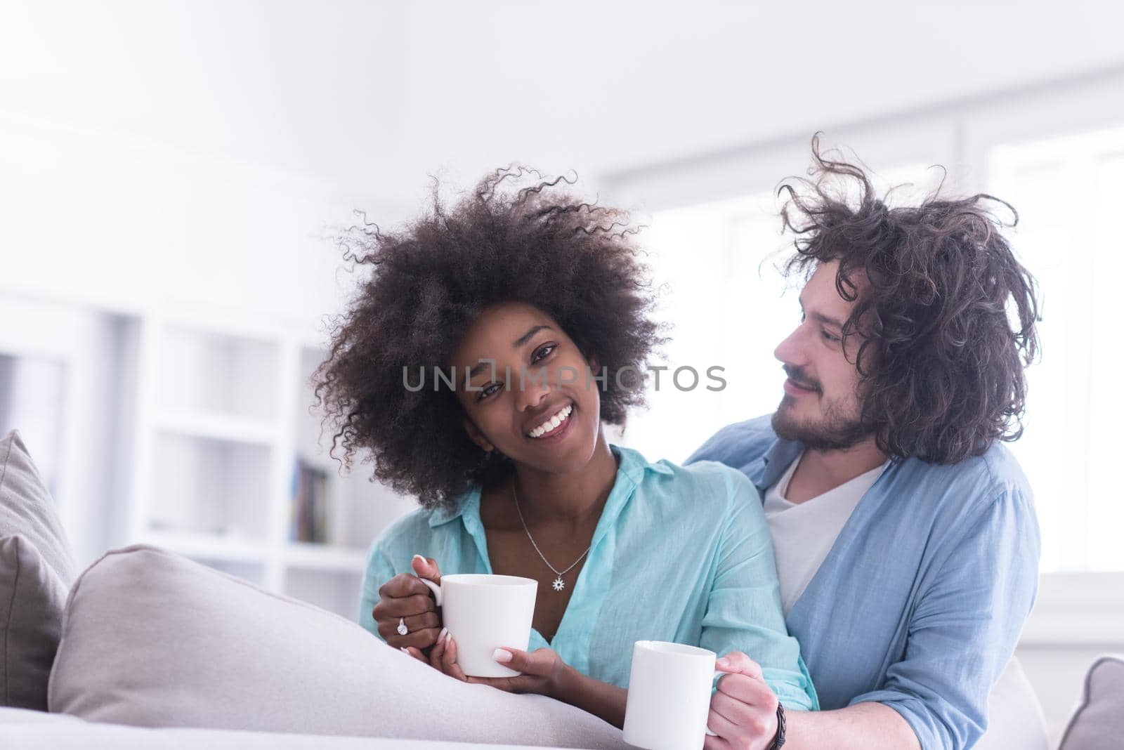 young multiethnic couple sitting on sofa at home drinking coffe, talking, smiling.