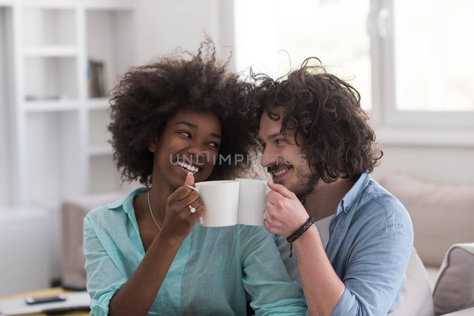 young multiethnic couple sitting on sofa at home drinking coffe, talking, smiling.