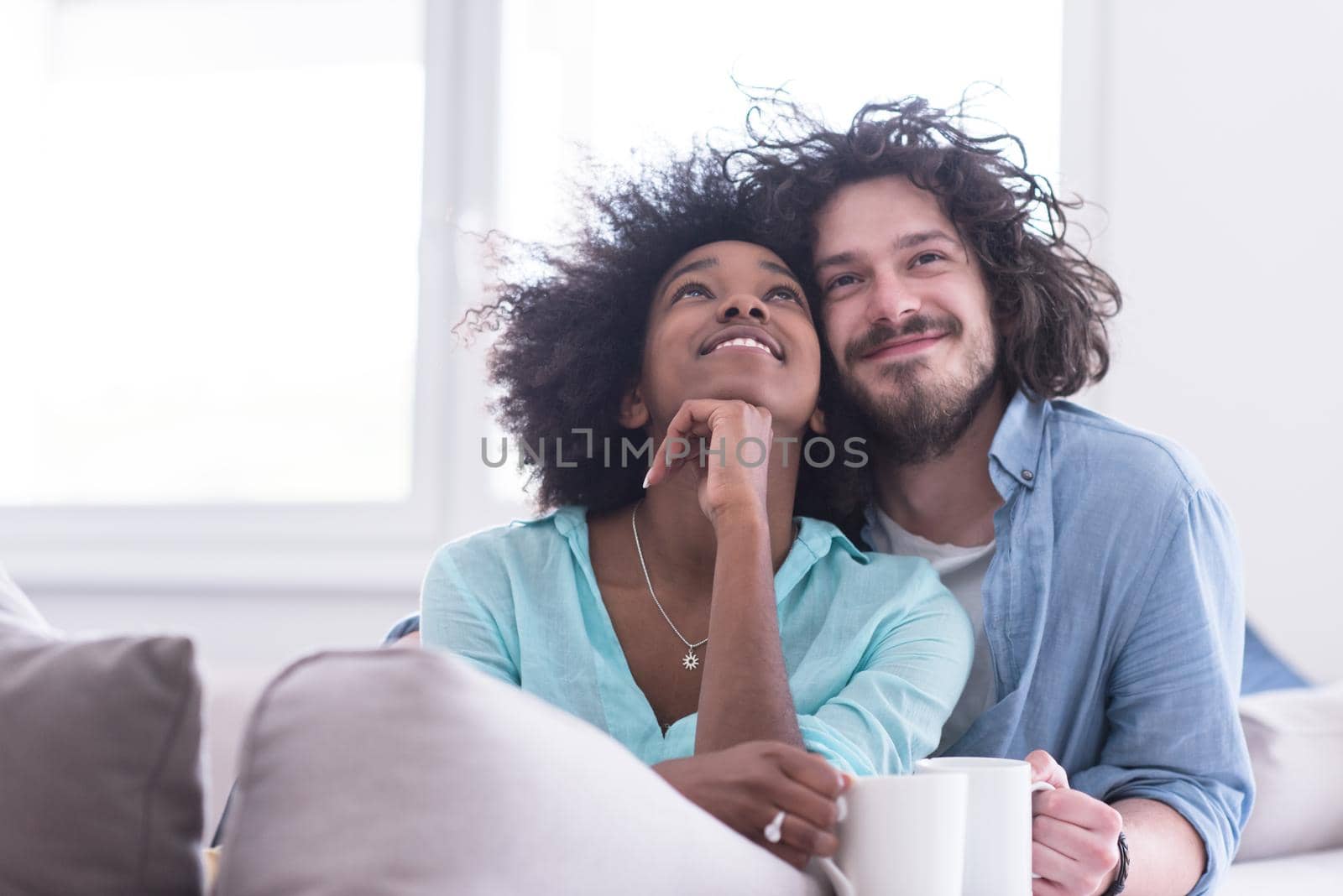 young multiethnic couple sitting on sofa at home drinking coffe, talking, smiling.