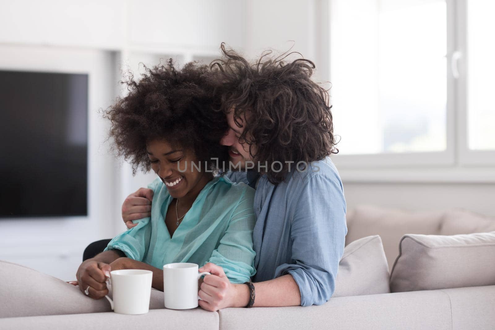 young multiethnic couple sitting on sofa at home drinking coffe, talking, smiling.