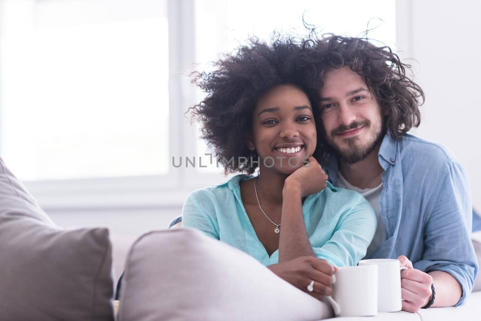 young multiethnic couple sitting on sofa at home drinking coffe, talking, smiling.