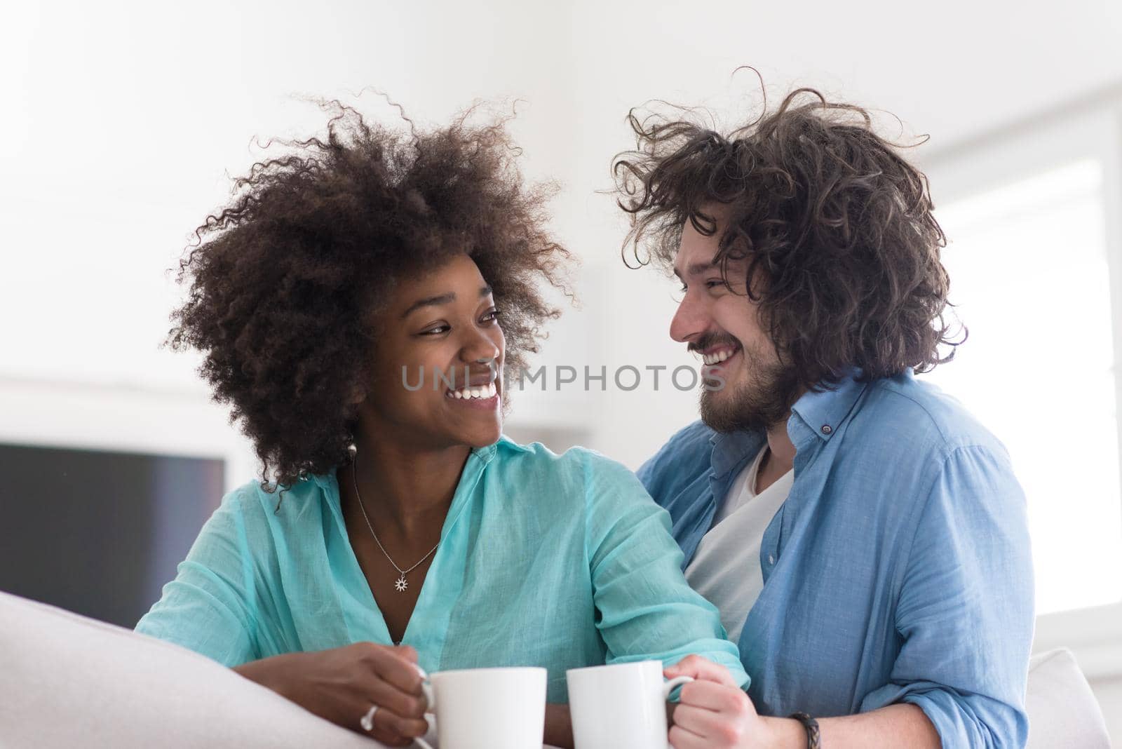 young multiethnic couple sitting on sofa at home drinking coffe, talking, smiling.