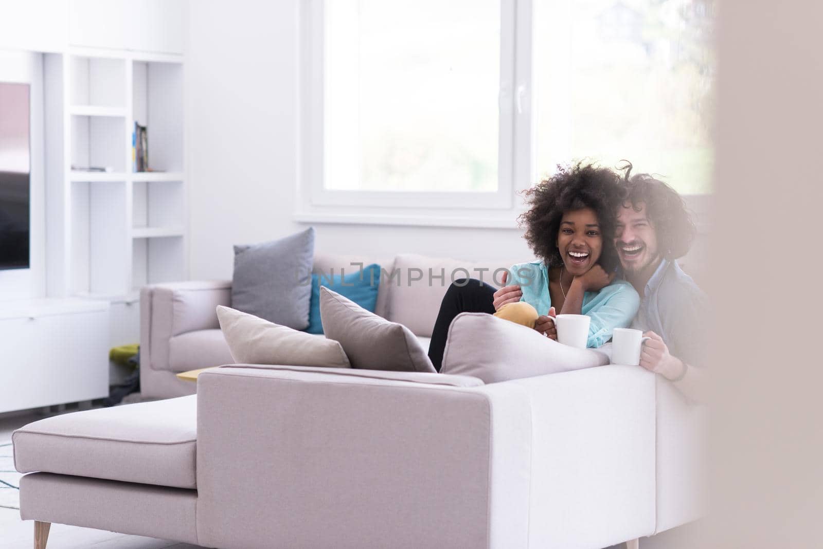 young multiethnic couple sitting on sofa at home drinking coffe, talking, smiling.