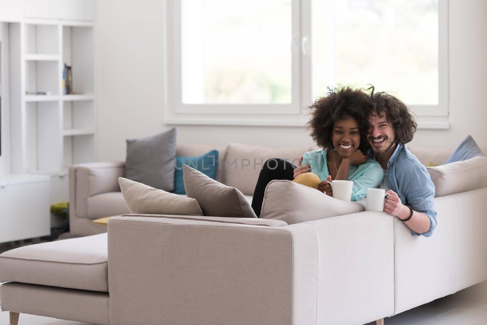 young multiethnic couple sitting on sofa at home drinking coffe, talking, smiling.