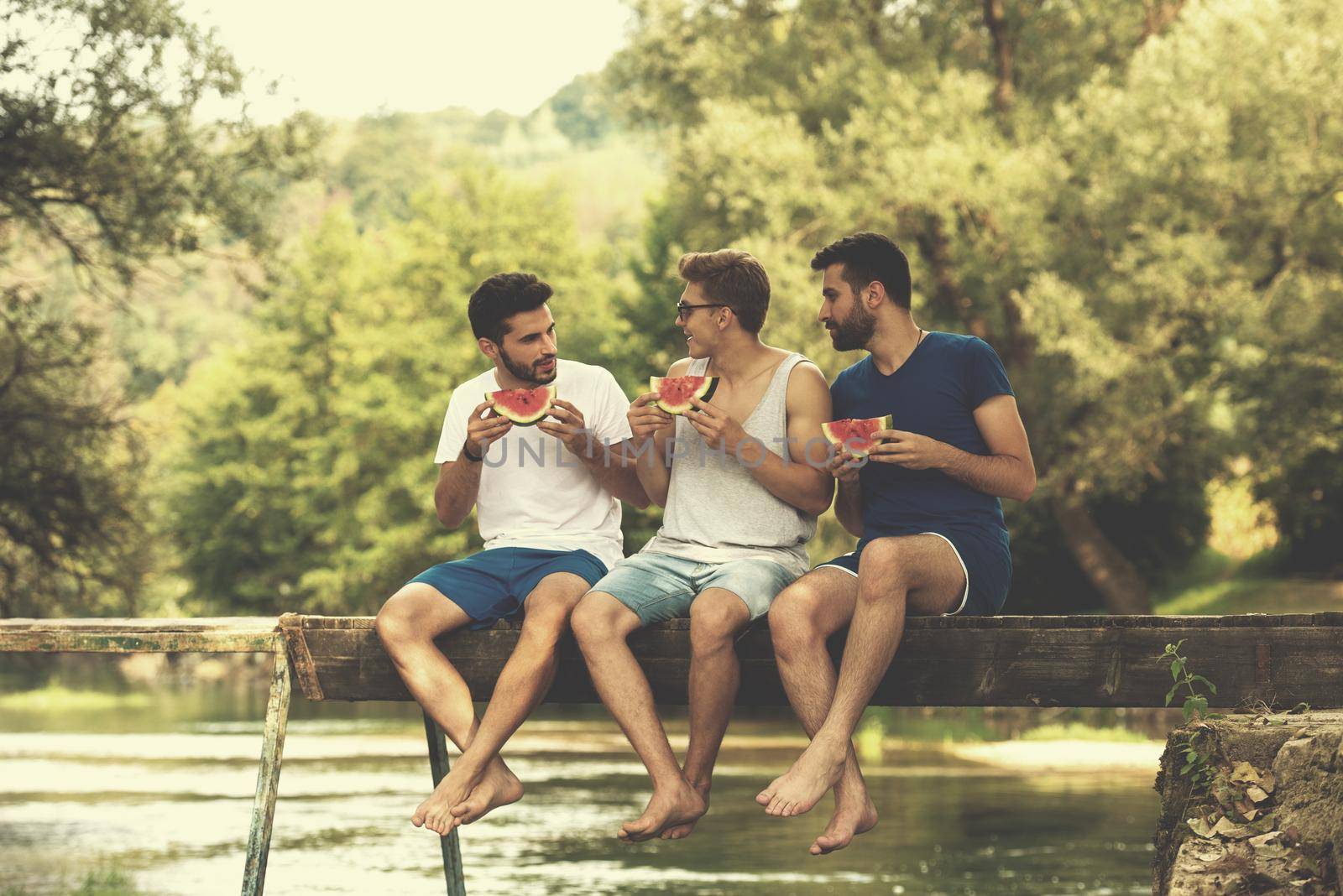 group of young men enjoying watermelon while sitting on the wooden bridge over the river in beautiful nature