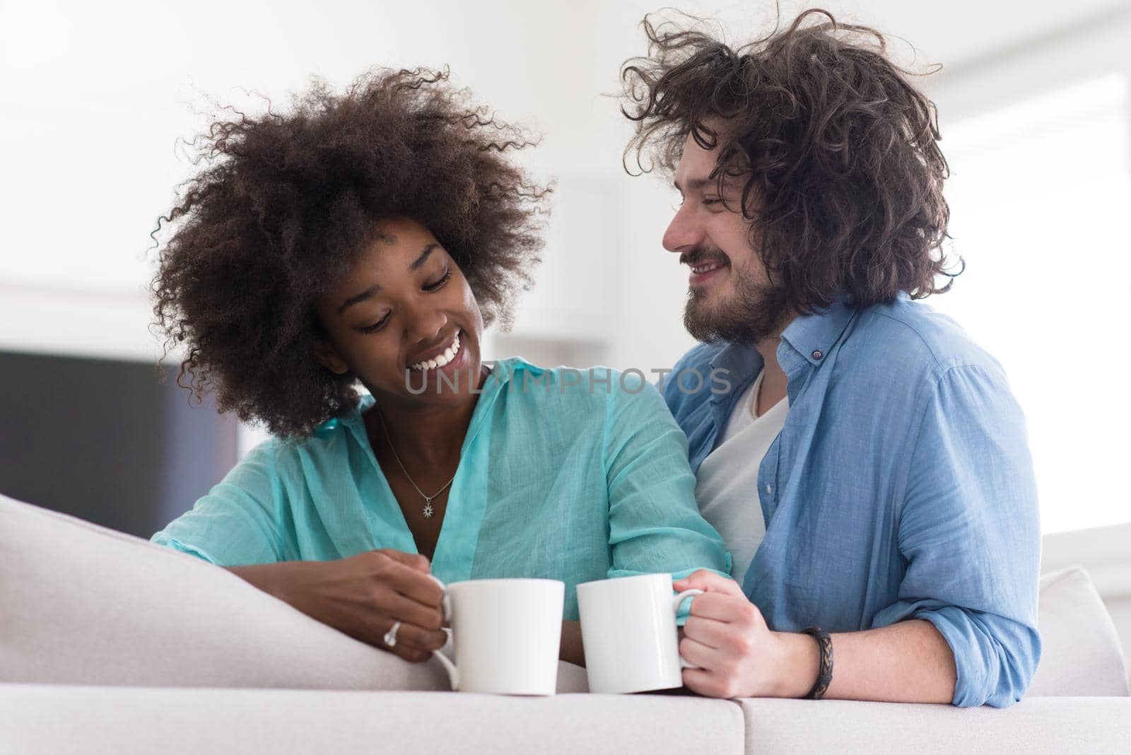 young multiethnic couple sitting on sofa at home drinking coffe, talking, smiling.