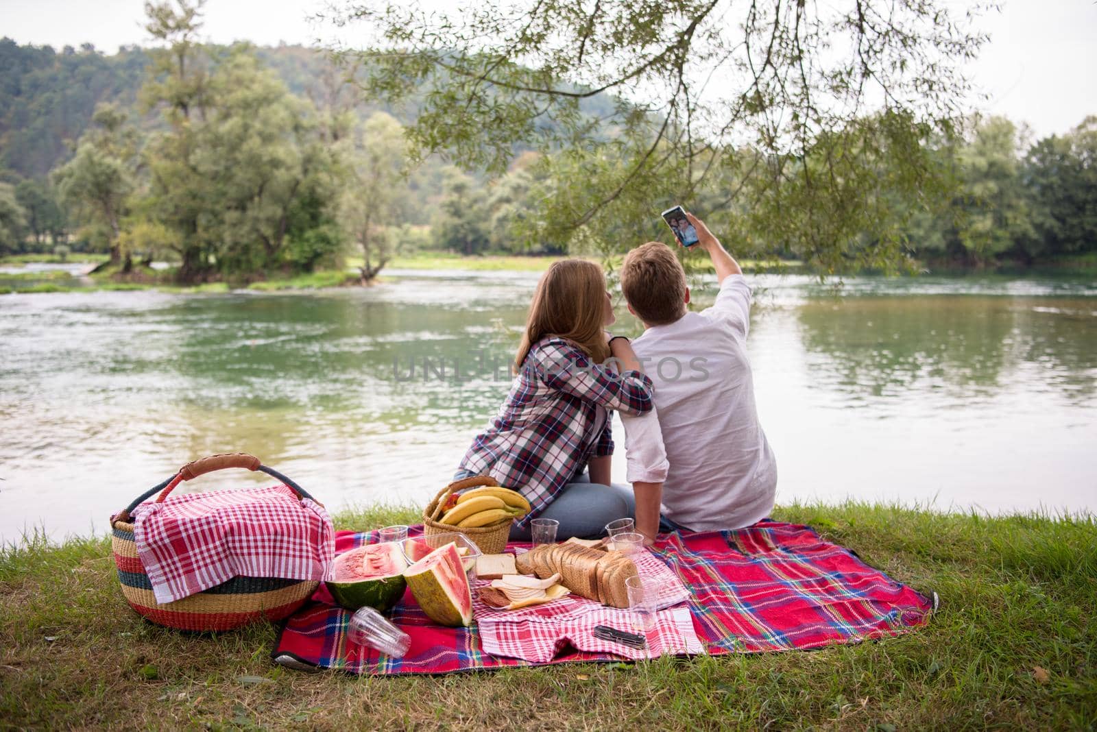 Couple in love taking a selfie by mobile phone while enjoying picnic time drink and food in beautiful nature on the river bank