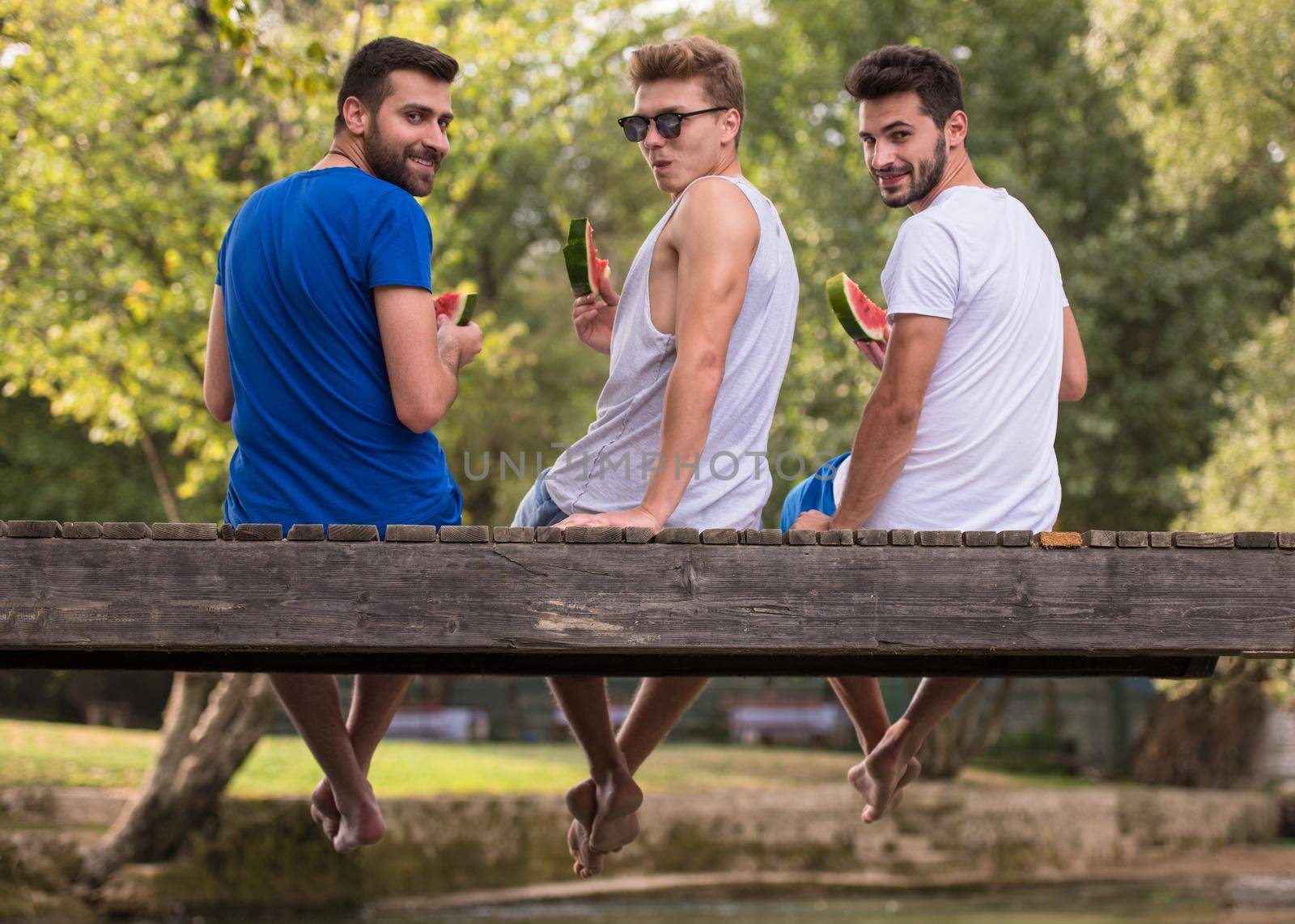 group of young men enjoying watermelon while sitting on the wooden bridge over the river in beautiful nature
