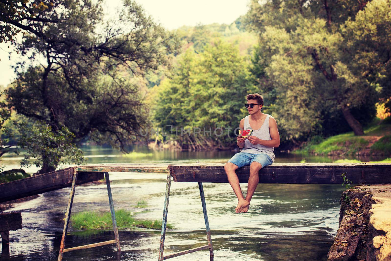young man enjoying watermelon while sitting on the wooden bridge over the river in beautiful nature
