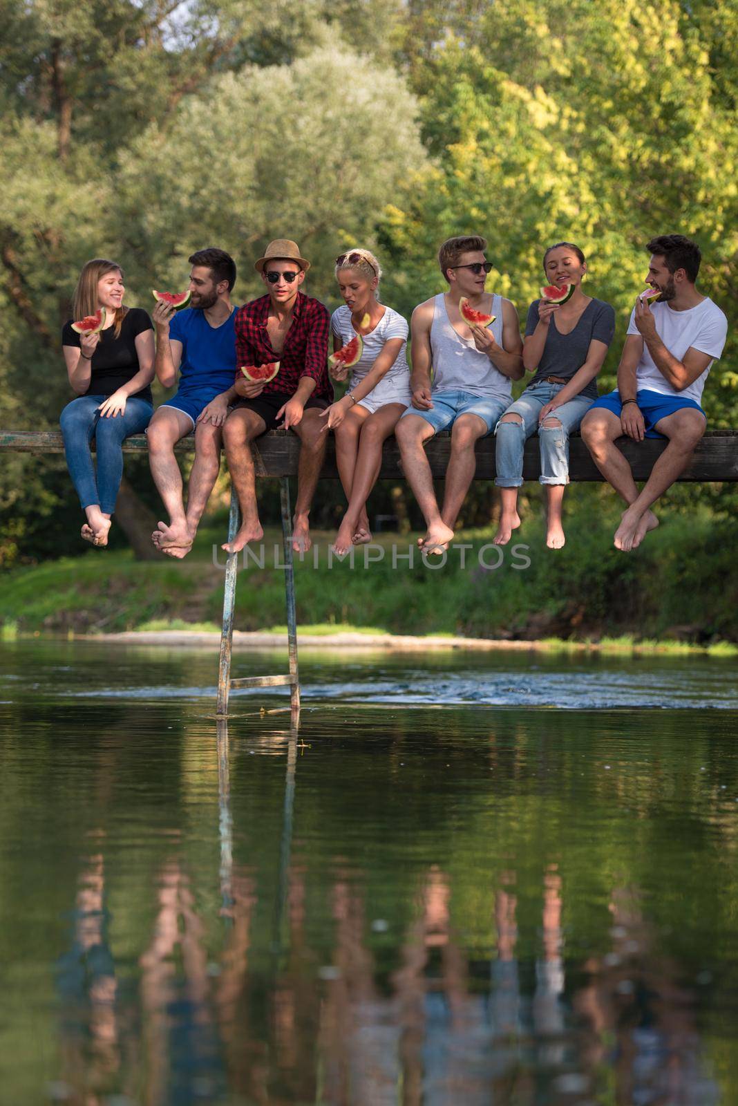 group of young friends enjoying watermelon while sitting on the wooden bridge over the river in beautiful nature