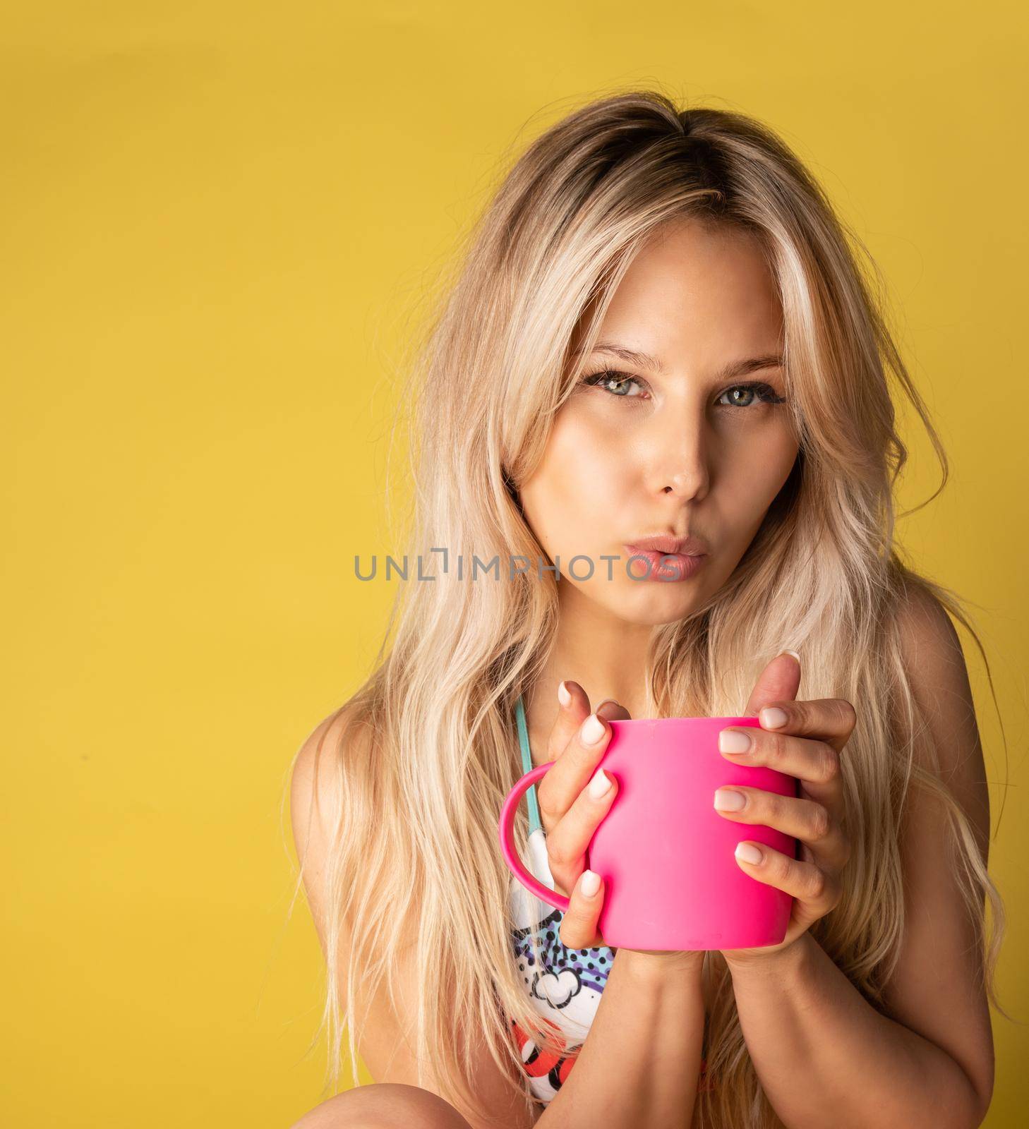 A girl sitting on the floor with her feet cross-legged keeps her head on a beautiful yellow background with a cup in hand yellow or pink and red bikini sexy, young female hair perfect, summer. Vacation adult lifestyle, caucasian soppy