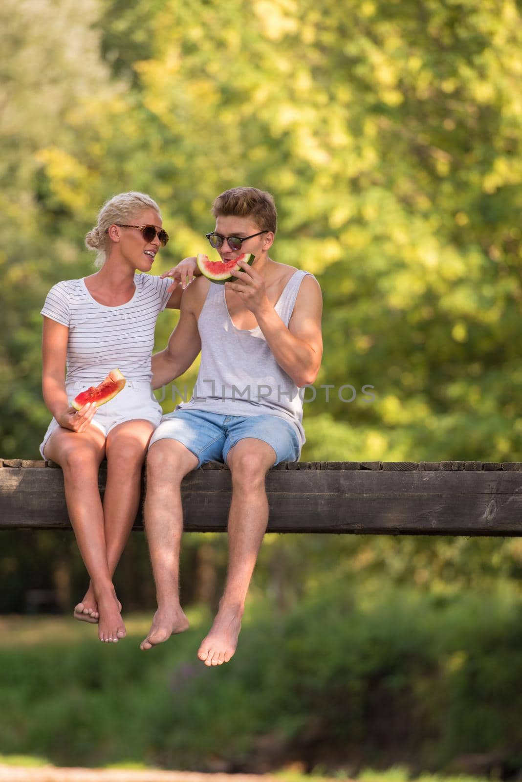 couple in love enjoying watermelon while sitting on the wooden bridge over the river in beautiful nature