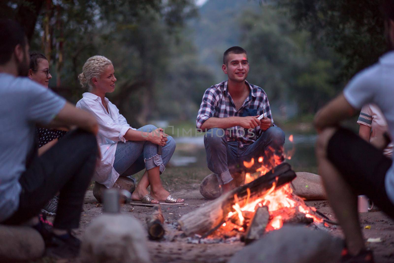 a group of happy young friends relaxing and enjoying  summer evening around campfire on the river bank