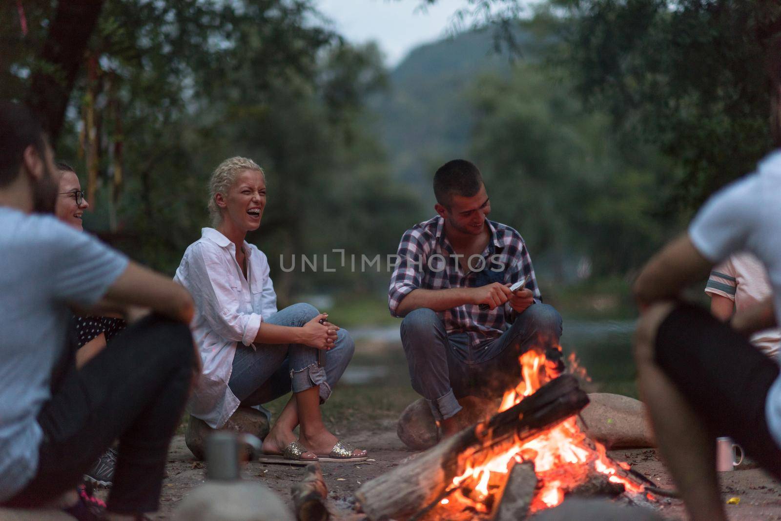 a group of happy young friends relaxing and enjoying  summer evening around campfire on the river bank