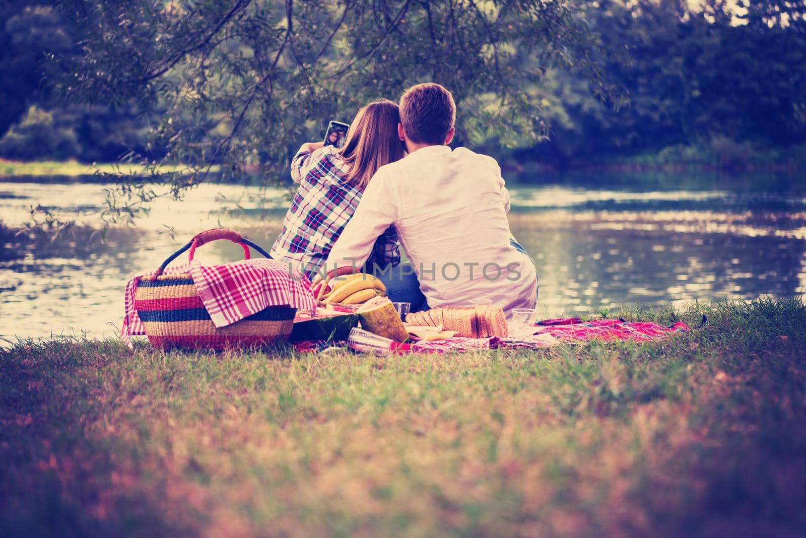 Couple in love taking a selfie by mobile phone while enjoying picnic time drink and food in beautiful nature on the river bank