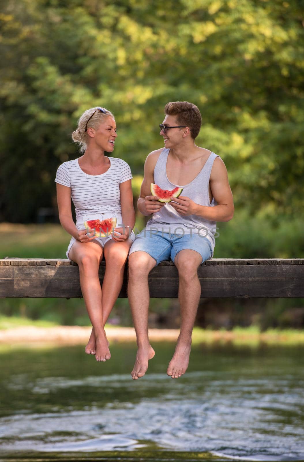 couple in love enjoying watermelon while sitting on the wooden bridge over the river in beautiful nature