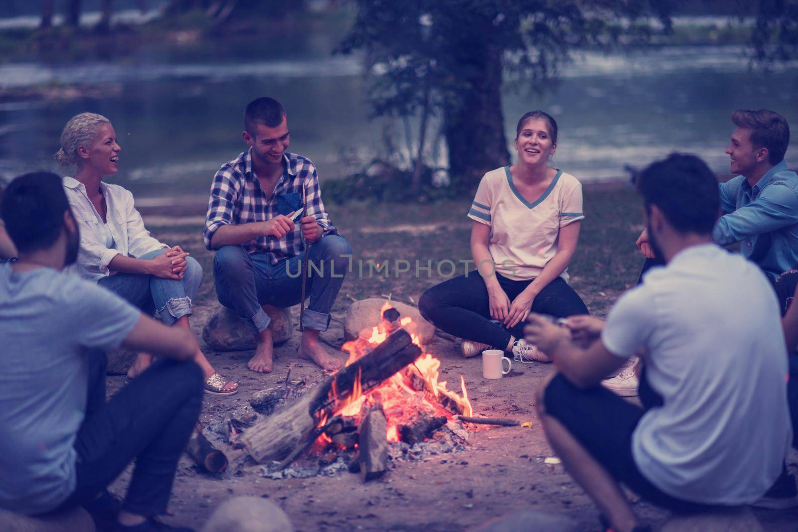 a group of happy young friends relaxing and enjoying  summer evening around campfire on the river bank